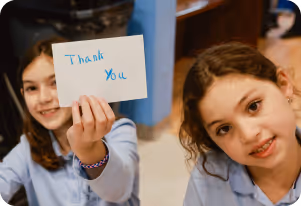 Two young girls in blue shirts smiling, one holding a card with the words 'Thank You'.