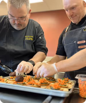 Two male chefs in black uniforms assembling appetizers with sliced vegetables on a tray in a kitchen.