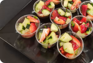 Small clear cups filled with diced cucumber and strawberry pieces, arranged on a black tray.