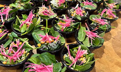 Multiple small bowls filled with fresh spinach leaves, shredded pink radish, and pine nuts on a wooden surface.
