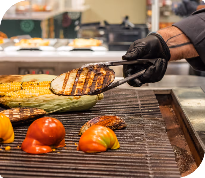 Grilled vegetables including corn on the cob, tomatoes, and eggplant being cooked on a grill with a gloved hand holding tongs.