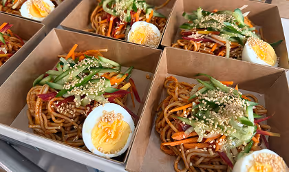 Takeout boxes with noodle salad topped with sliced cucumber, shredded carrots, sesame seeds, and halved boiled eggs.
