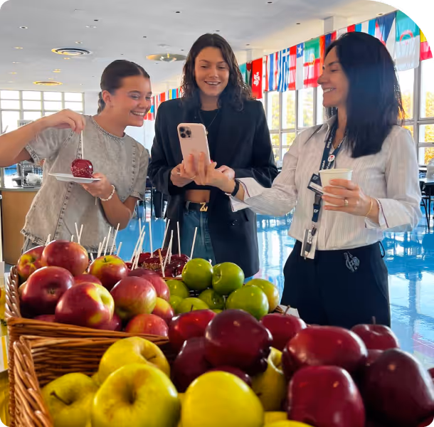 Three women looking at a smartphone and smiling near baskets of red, green, and yellow apples indoors with international flags in the background.