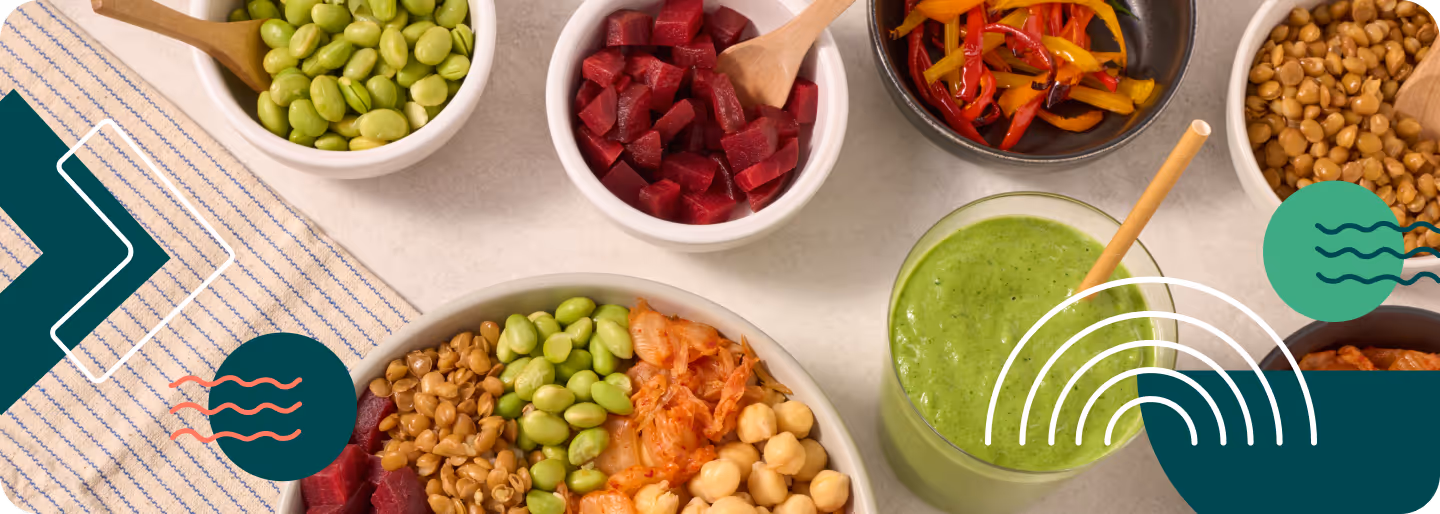 Assortment of bowls containing chickpeas, lentils, edamame, diced beets, roasted peppers, kimchi, and a green smoothie with a straw on a light surface.