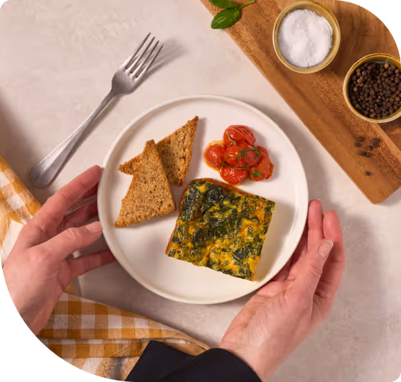 Hands holding a plate with spinach and egg frittata, roasted cherry tomatoes, and two pieces of toasted bread, with a fork and bowls of salt and pepper on the side.
