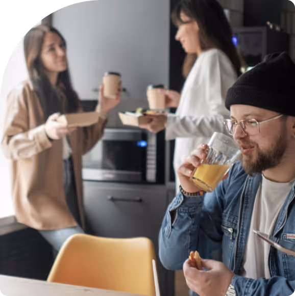 Man wearing glasses and a beanie drinking orange juice while holding a sandwich, with two women in the background holding food containers and coffee cups in a kitchen setting.