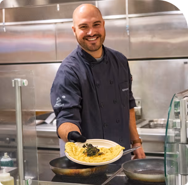 Smiling chef holding a plate with an omelette garnished with herbs in a professional kitchen.