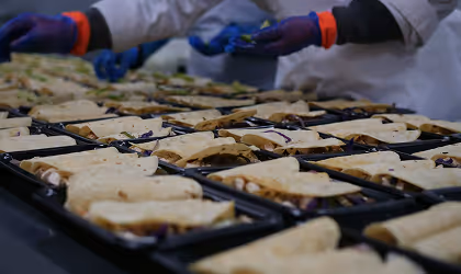 Factory worker wearing gloves assembling sandwiches in a production line with multiple trays of sandwiches.