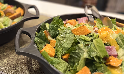 Close-up of a fresh salad with leafy greens, roasted sweet potato chunks, and other vegetables in a black serving dish with tongs.