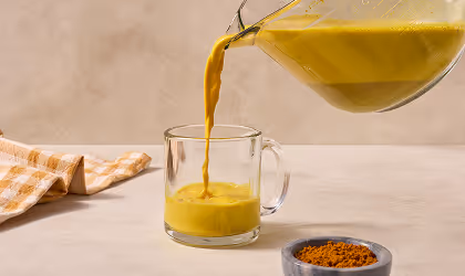 Golden turmeric latte being poured from a glass pitcher into a clear mug, next to a bowl of turmeric powder and a checkered cloth.