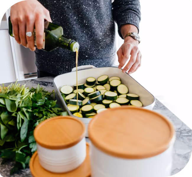 Person pouring olive oil over sliced zucchini in a baking dish next to fresh greens on a countertop.