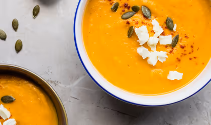 Two bowls of creamy orange soup, one topped with pumpkin seeds and crumbled cheese, on a light gray surface.