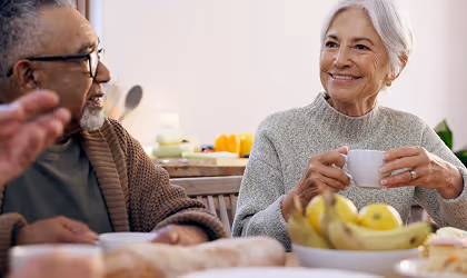 Smiling elderly woman holding a white cup and conversing with an elderly man at a kitchen table with fruit and bread.