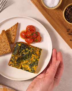 Plate with two slices of toasted bread, a square piece of spinach and egg frittata, and roasted cherry tomatoes, with a hand reaching toward the plate.