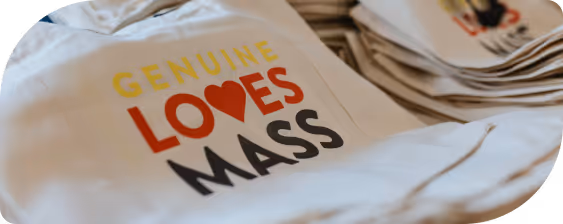 Close-up of a beige tote bag with the text 'GENUINE LOVES MASS' printed, where the letter 'O' is replaced by a red heart.