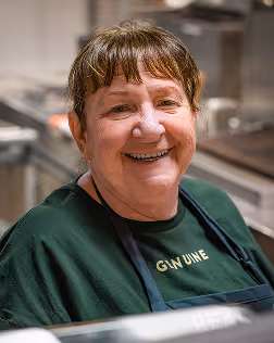 Smiling older woman with short hair wearing an apron in a kitchen setting.