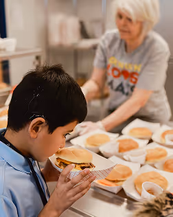 Young boy with a hearing aid smelling a hamburger in a cafeteria with a woman serving food in the background.