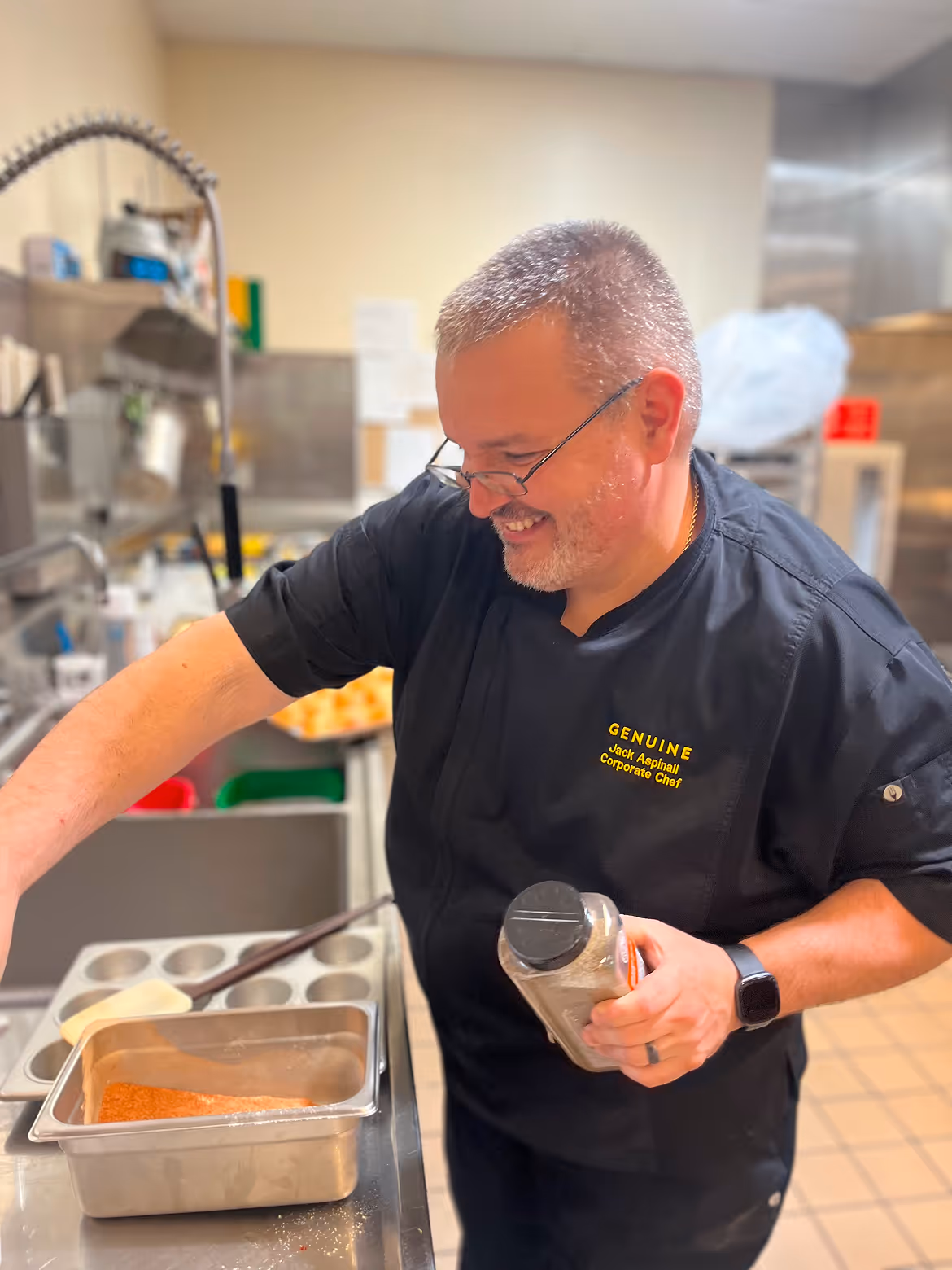 Smiling chef in a black uniform seasoning food in a stainless steel kitchen.