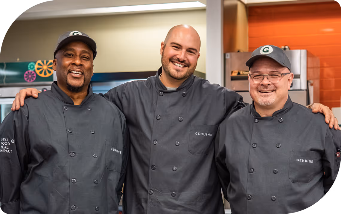 Three chefs in gray uniforms smiling, standing side by side with arms around each other in a kitchen.