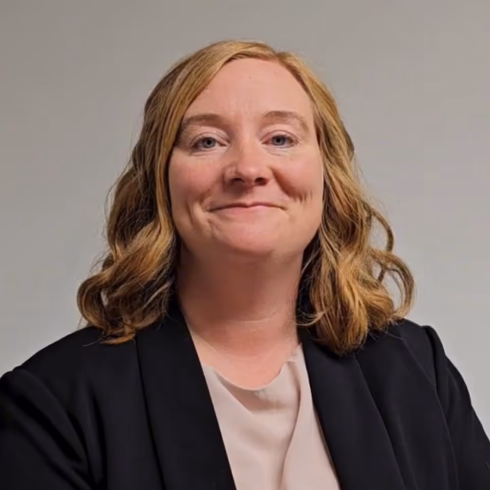 Smiling woman with shoulder-length wavy blonde hair wearing a black blazer over a beige blouse against a plain gray background.