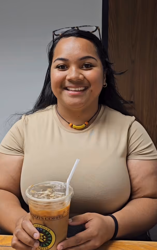 Smiling woman with long dark hair and glasses on her head holds a plastic cup of iced coffee with a white straw indoors.