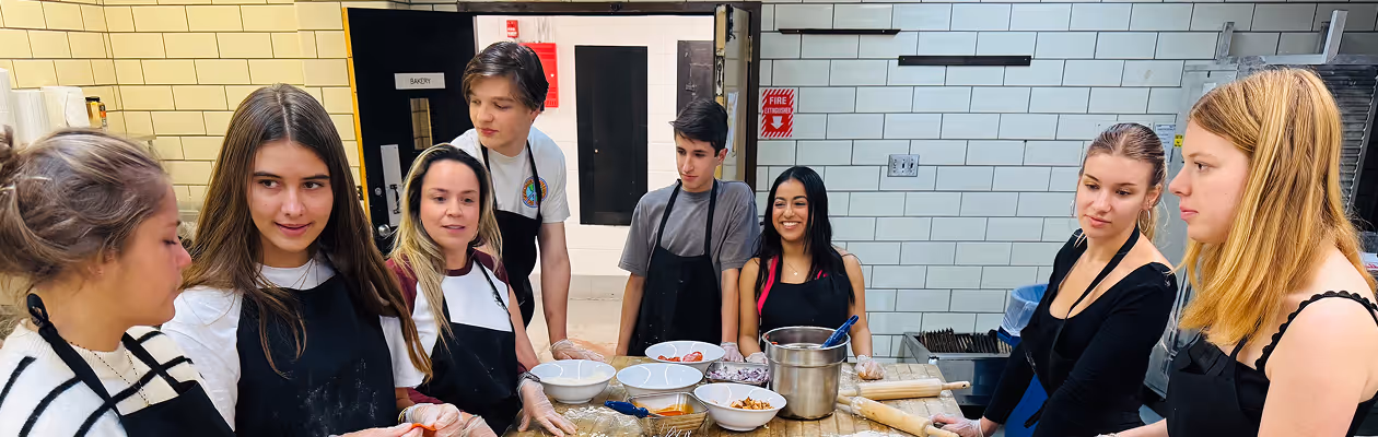 Seven young adults wearing aprons standing around a wooden table in a kitchen with bowls of ingredients and rolling pins.