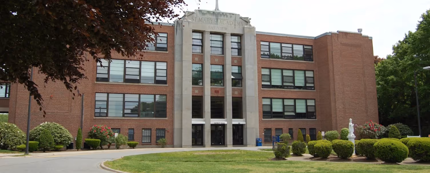 Front view of St. Mary’s Academy Bay View building with brick facade, large windows, and a lawn with trimmed bushes and a statue.