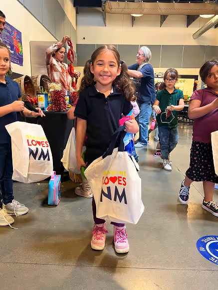 Smiling young girl standing indoors holding a tote bag that says 'New Mexico Loves NM' with other children and adults in the background.