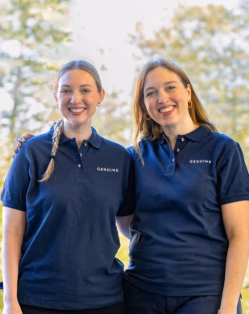 Two smiling women wearing navy polo shirts with the word 'GENUINE' embroidered, standing close together outdoors.
