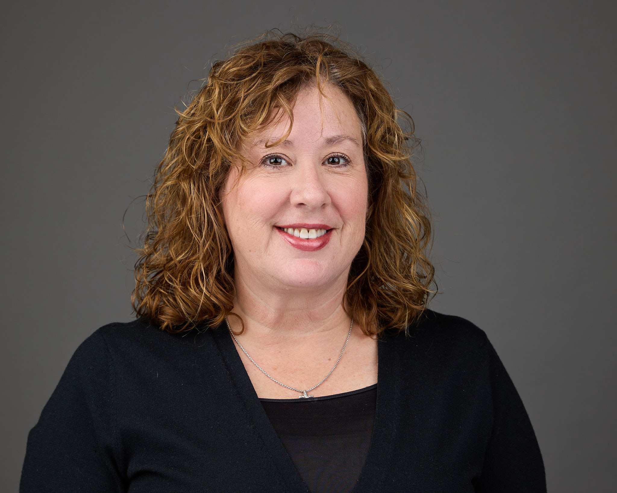 Smiling woman with curly light brown hair wearing a black top and silver necklace against a gray background.