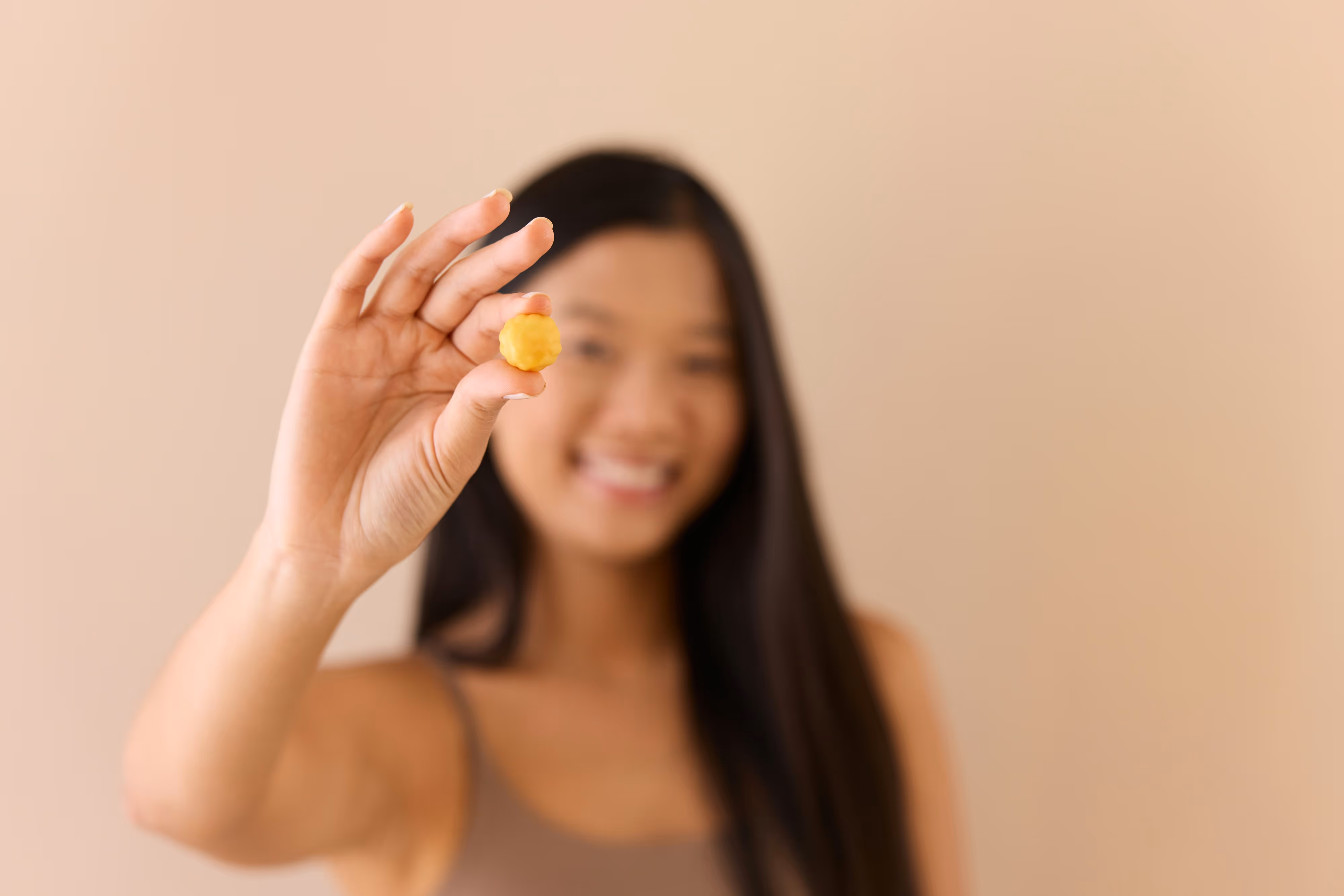 Une femme souriante tenant une petite boule jaune entre le pouce et l'index devant un fond beige flou.