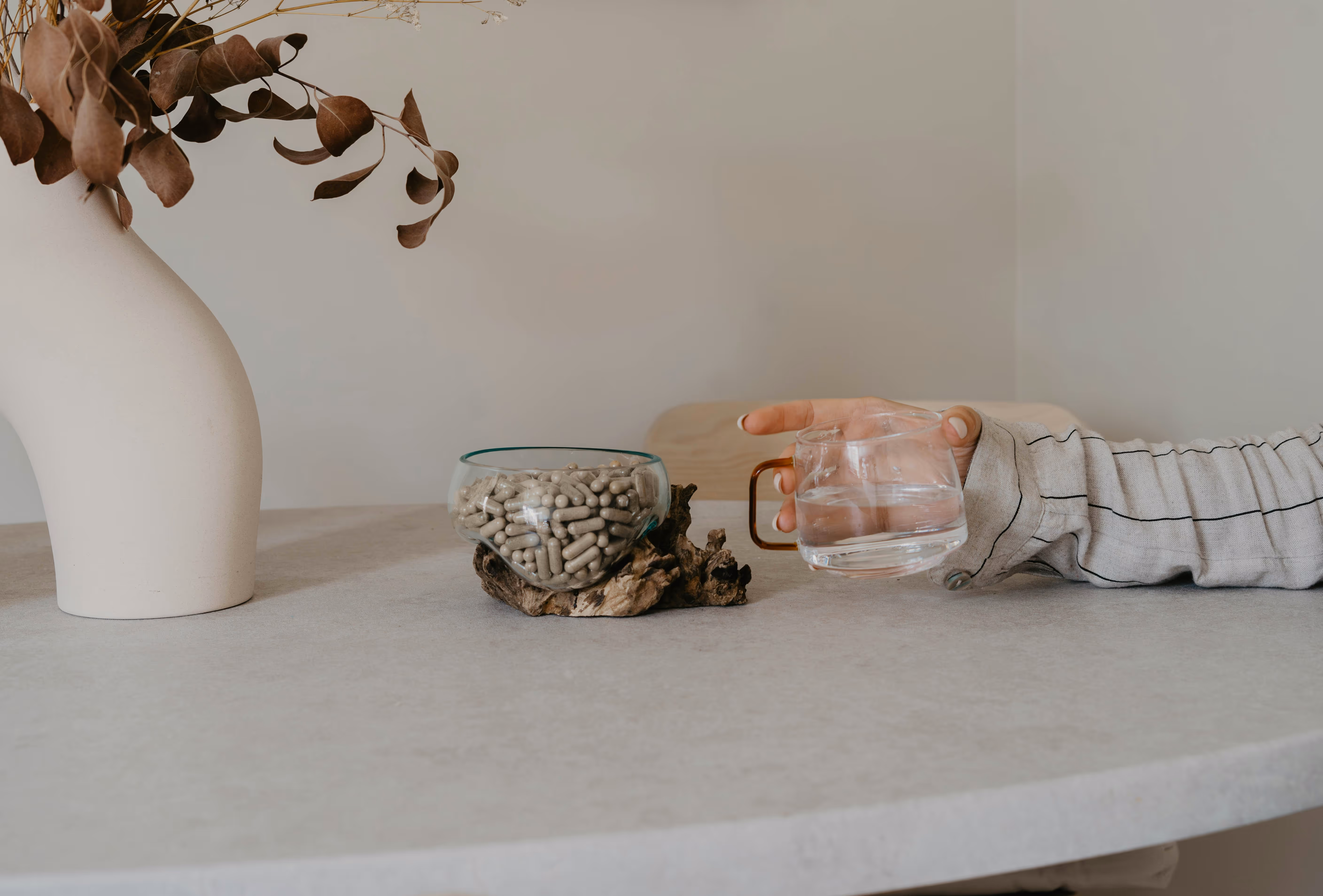 Main tenant un verre d'eau près d'un bol en verre contenant des capsules, posé sur une table avec un vase décoratif à côté.