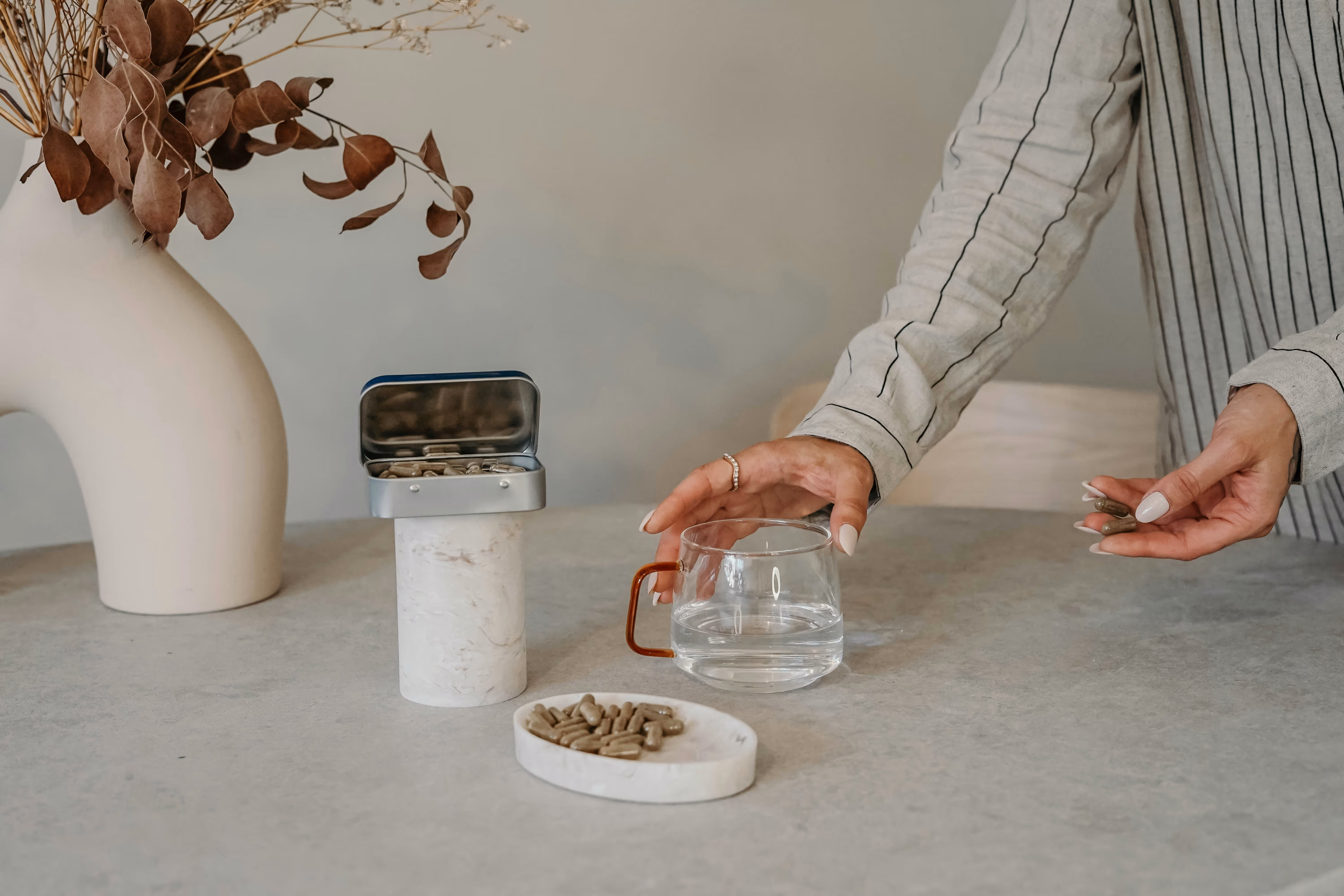 Une main tenant deux capsules près d'un verre d'eau sur une table avec une boîte et un plateau contenant des capsules, à côté d'un vase avec des feuilles séchées.