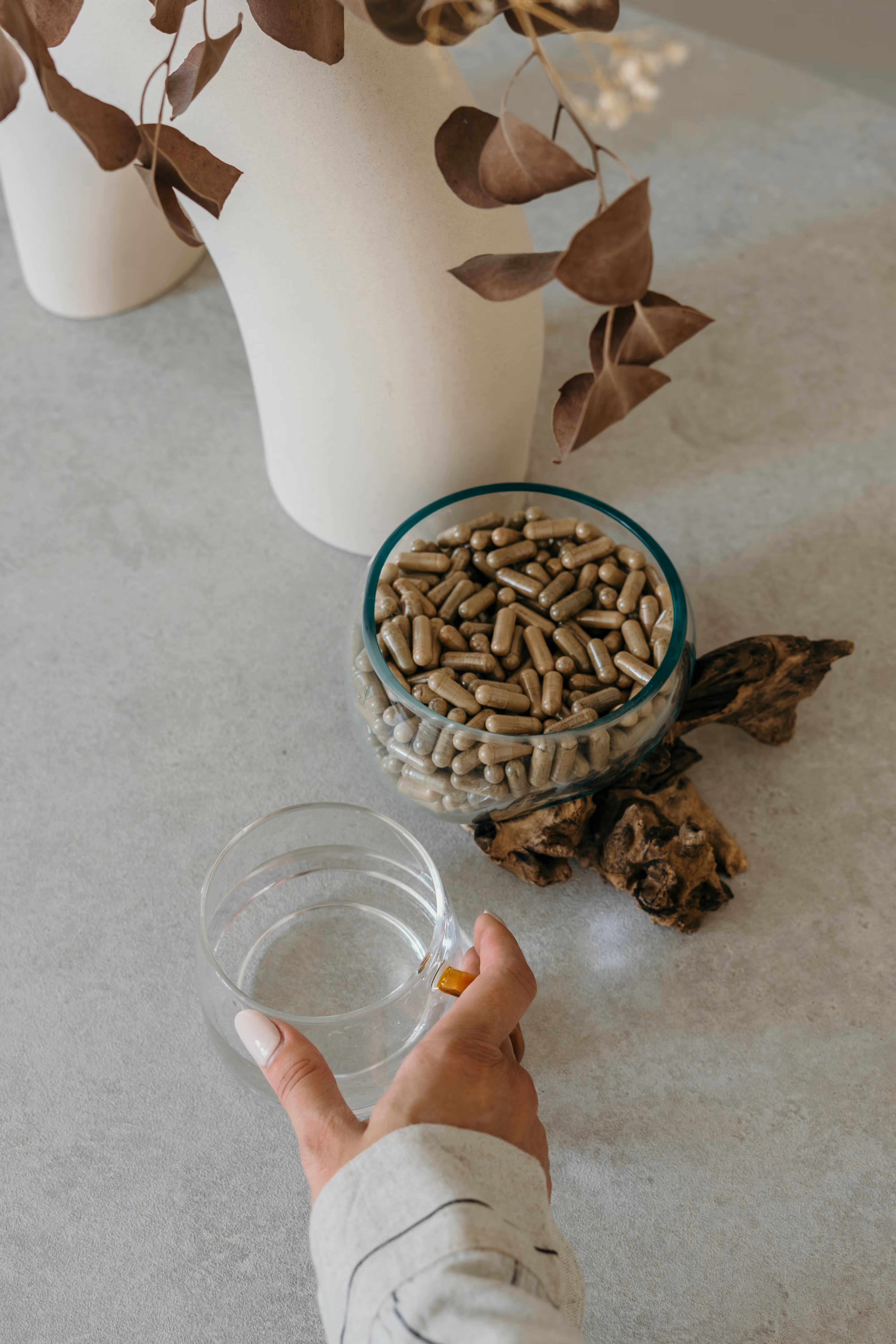 Main tenant une tasse en verre d'eau à côté d'un bol en verre rempli de capsules sur une table grise, avec un pot beige et des feuilles séchées en arrière-plan.