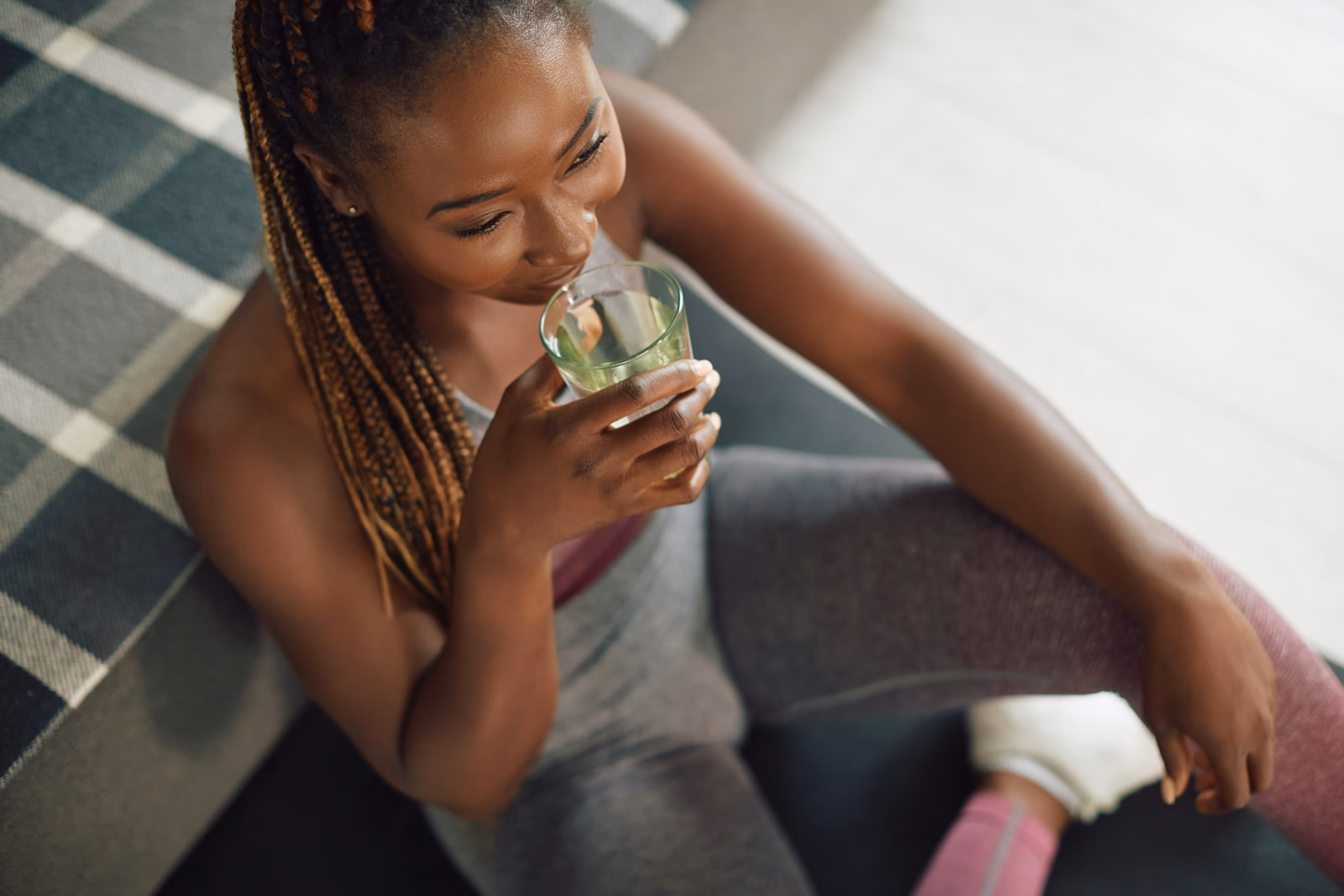 Femme assise sur un tapis à carreaux, portant des vêtements de sport, buvant dans un verre transparent.