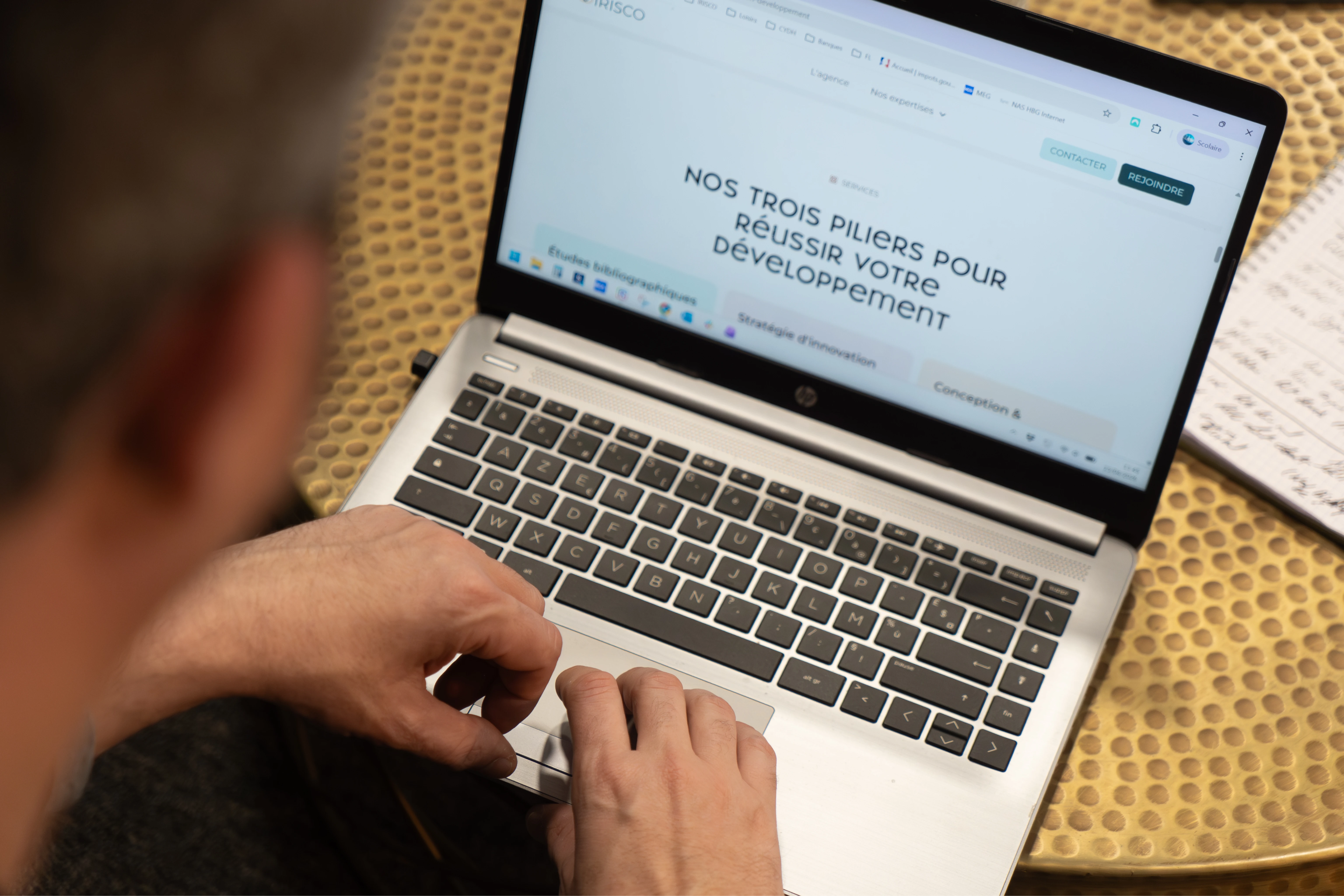 Personne utilisant un ordinateur portable HP affichant la page intitulée « Nos trois piliers pour réussir votre développement » sur une table en métal texturé.