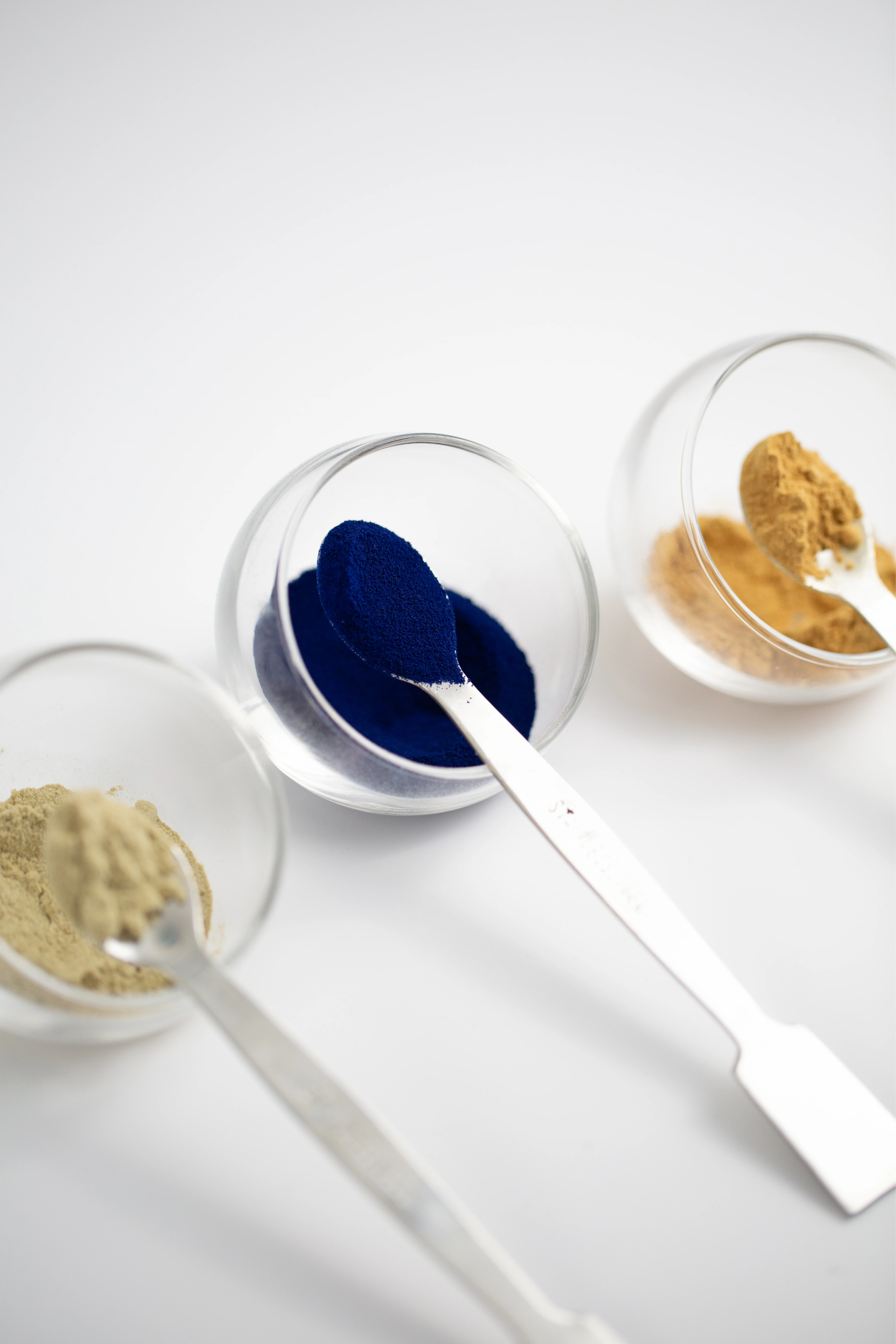 Trois cuillères remplies de poudres de différentes couleurs disposées dans des petits bols en verre sur fond blanc.