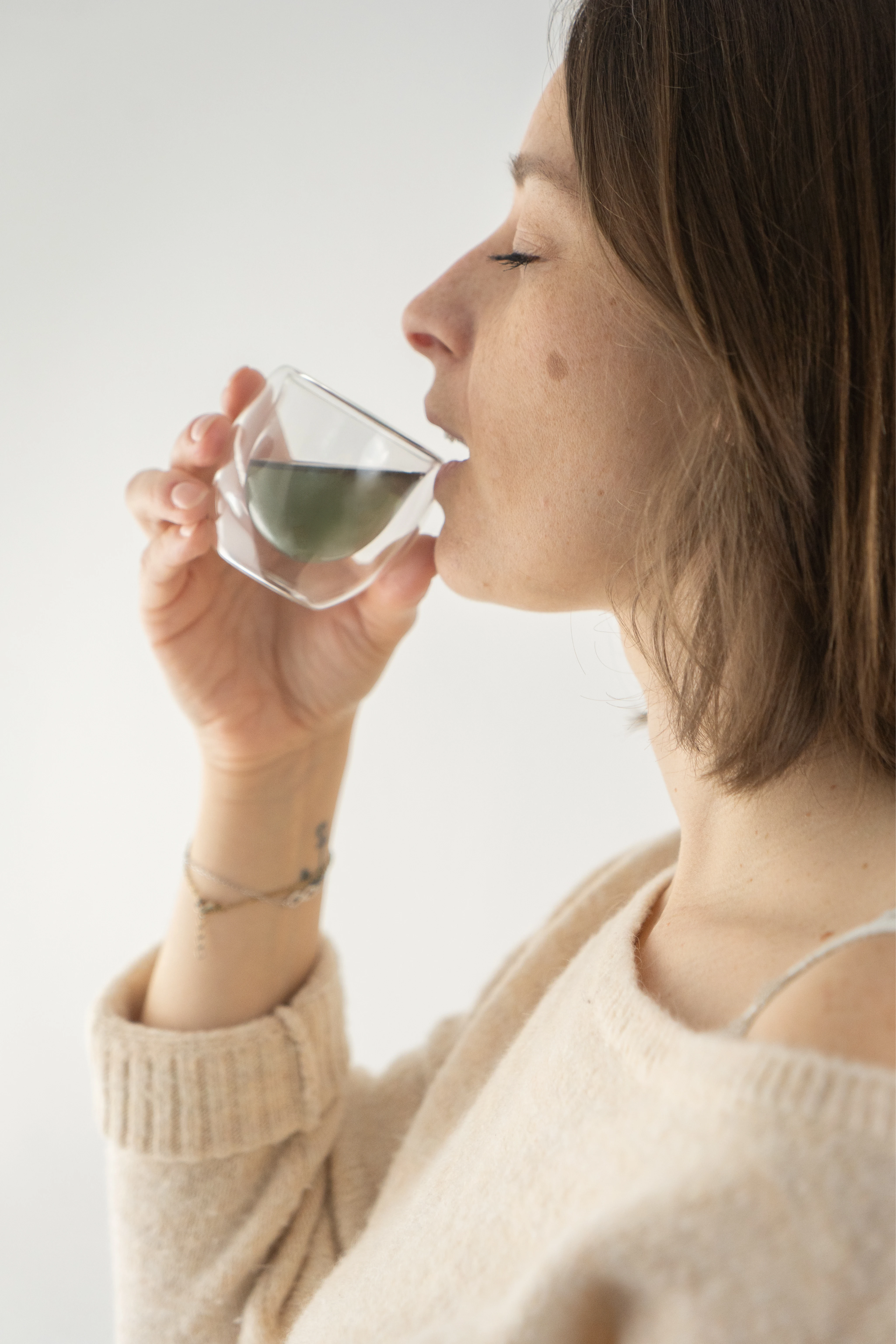 Femme buvant une boisson chaude verte dans une tasse en verre transparent, portant un pull beige.