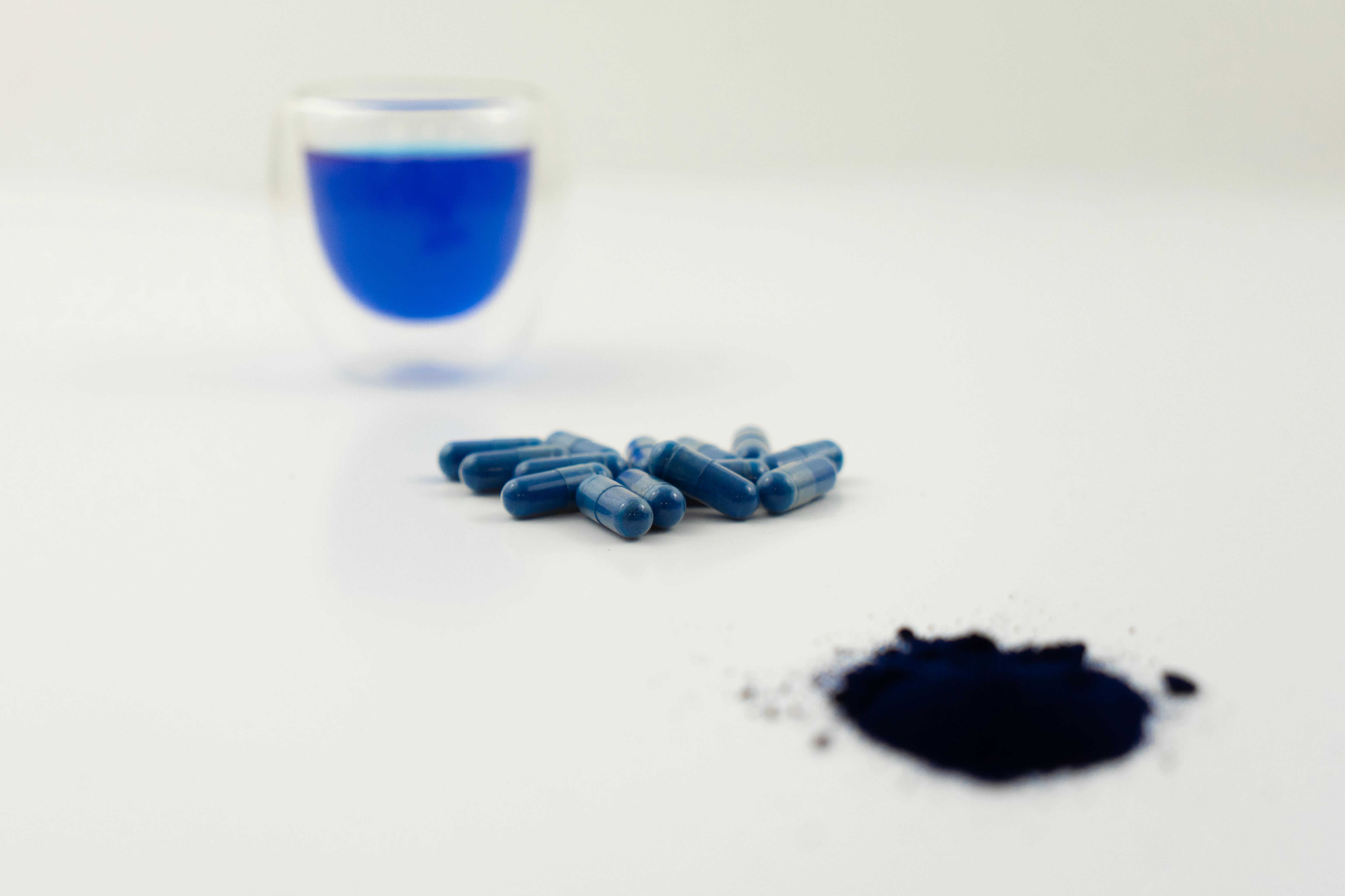 Capsules bleues, poudre noire et verre contenant un liquide bleu sur une surface blanche.