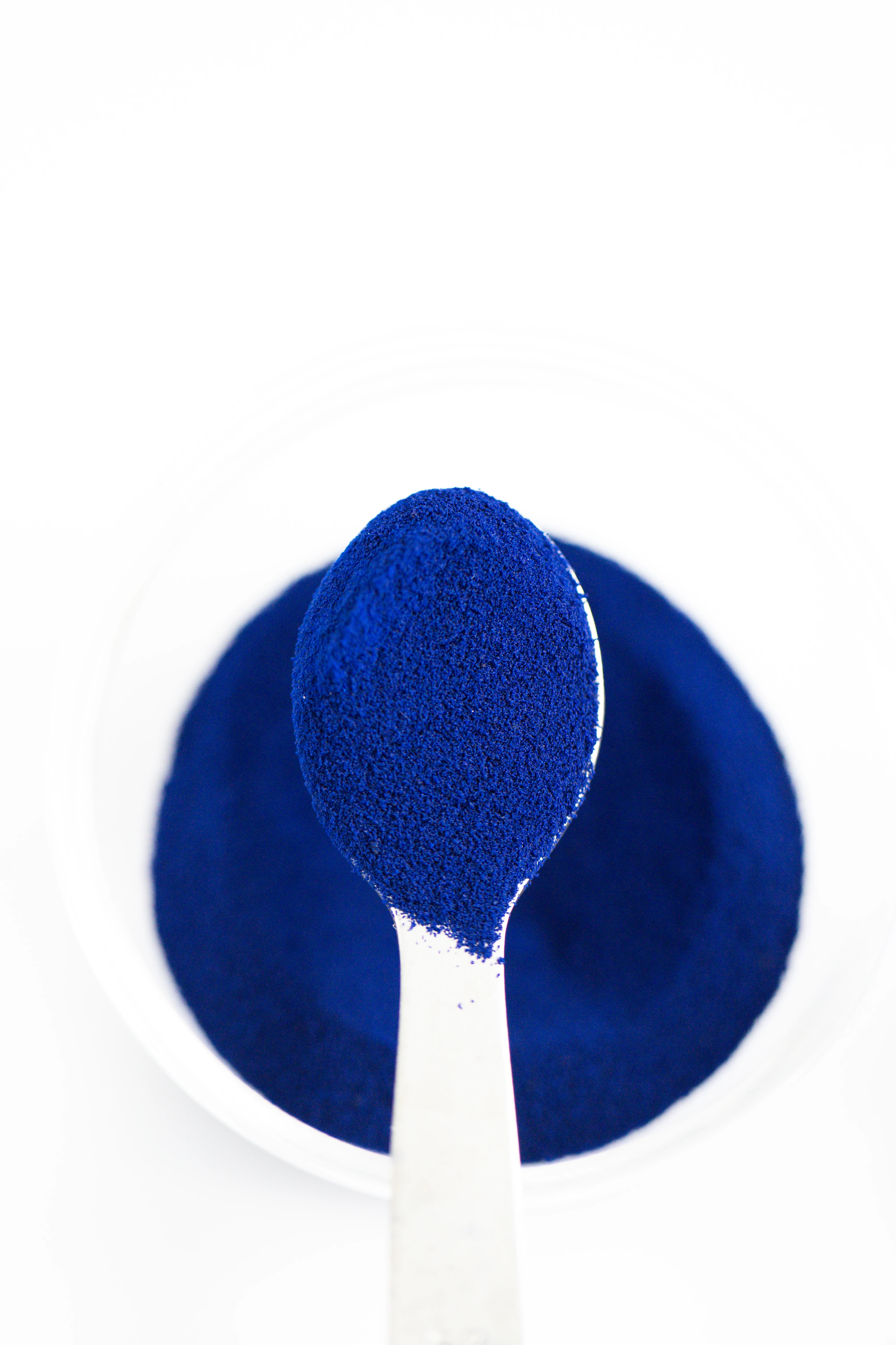 Cuillère remplie de poudre bleu vif placée au-dessus d'un bol blanc contenant plus de la même poudre.