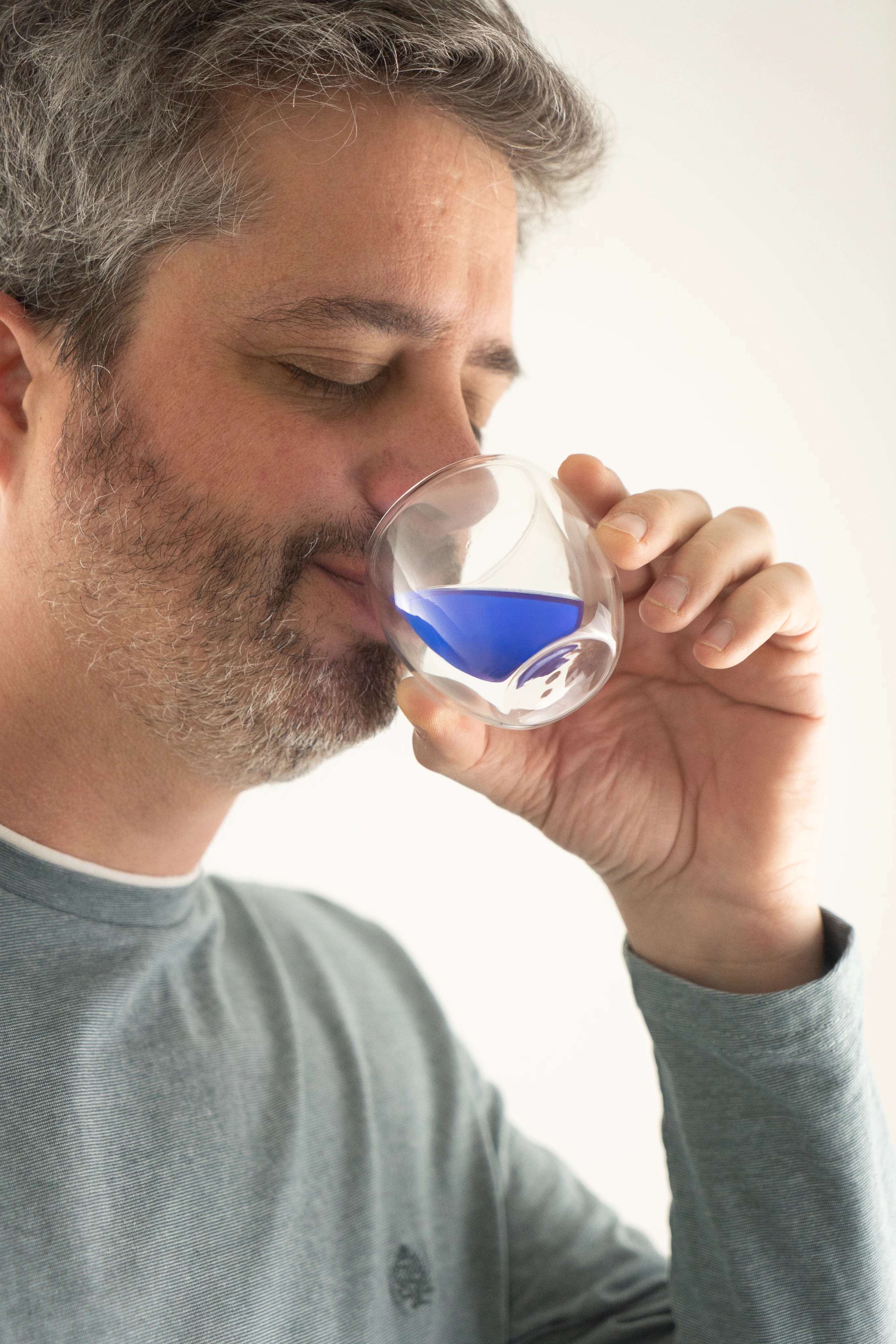 Homme moustachu portant un t-shirt gris, buvant dans un petit verre transparent contenant un liquide bleu.