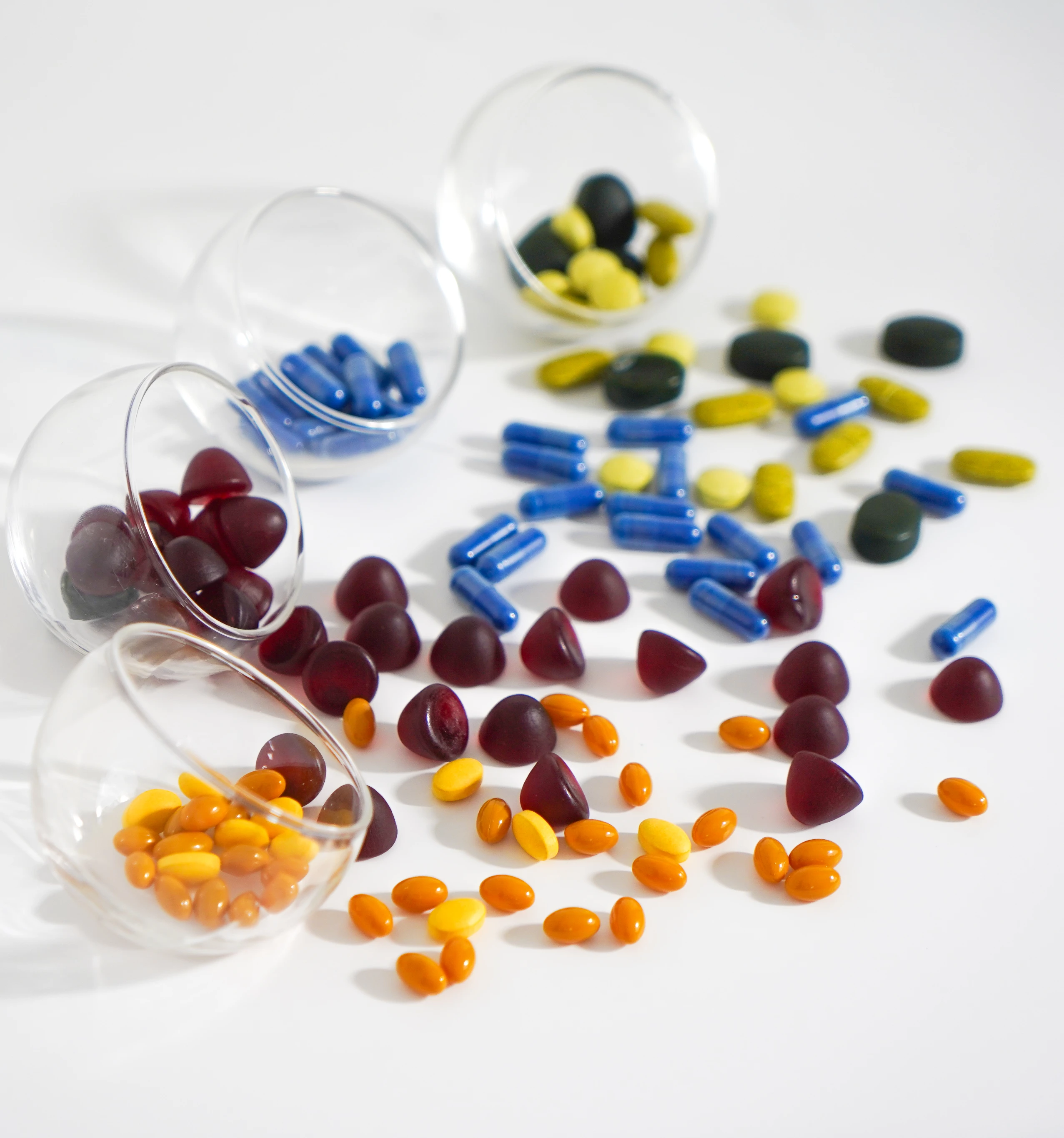 Différents types de comprimés et capsules colorés déversés à partir de quatre petits contenants en verre transparents sur une surface blanche.