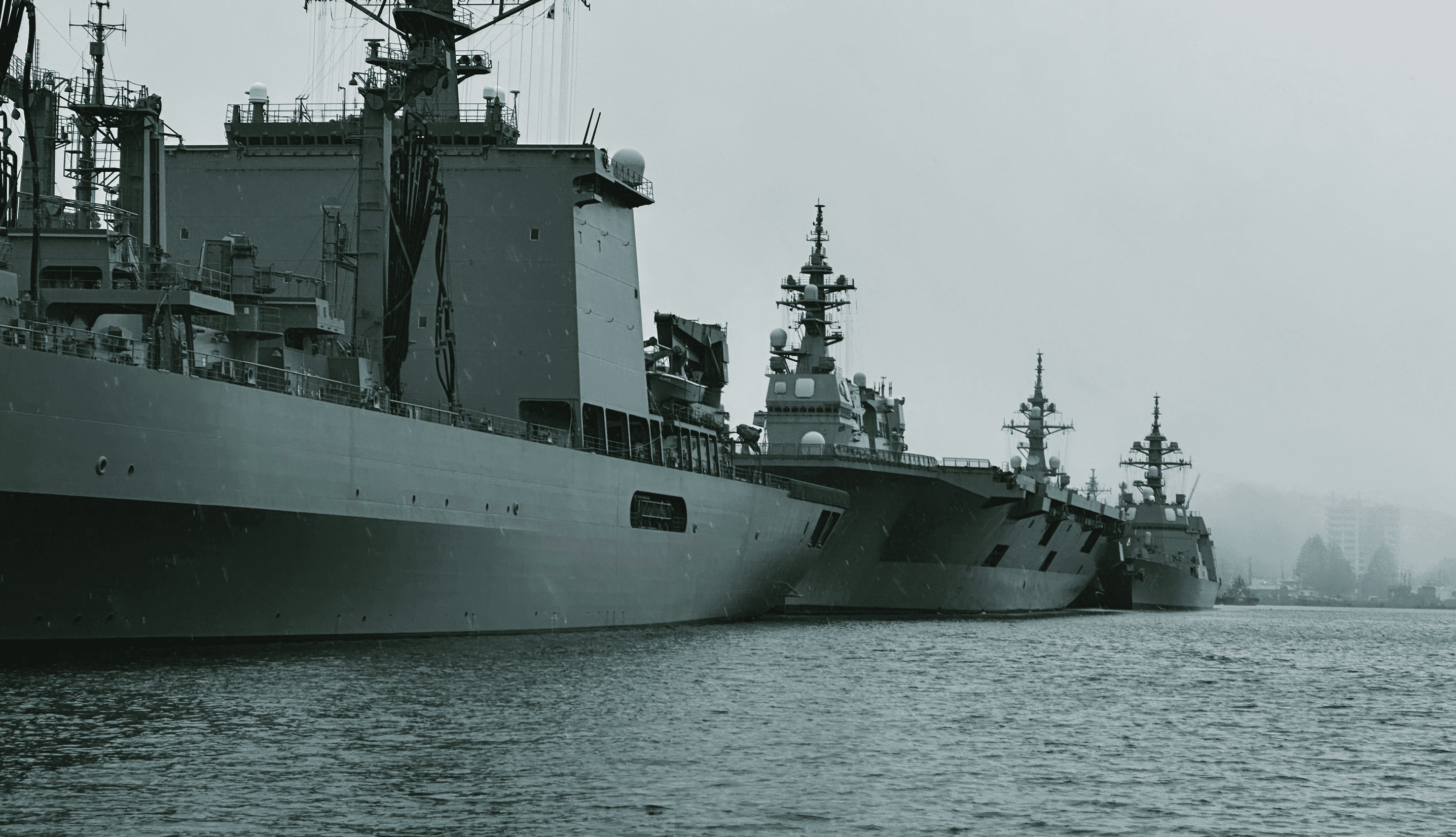 Several large naval warships docked side by side on a calm body of water under a gray sky.