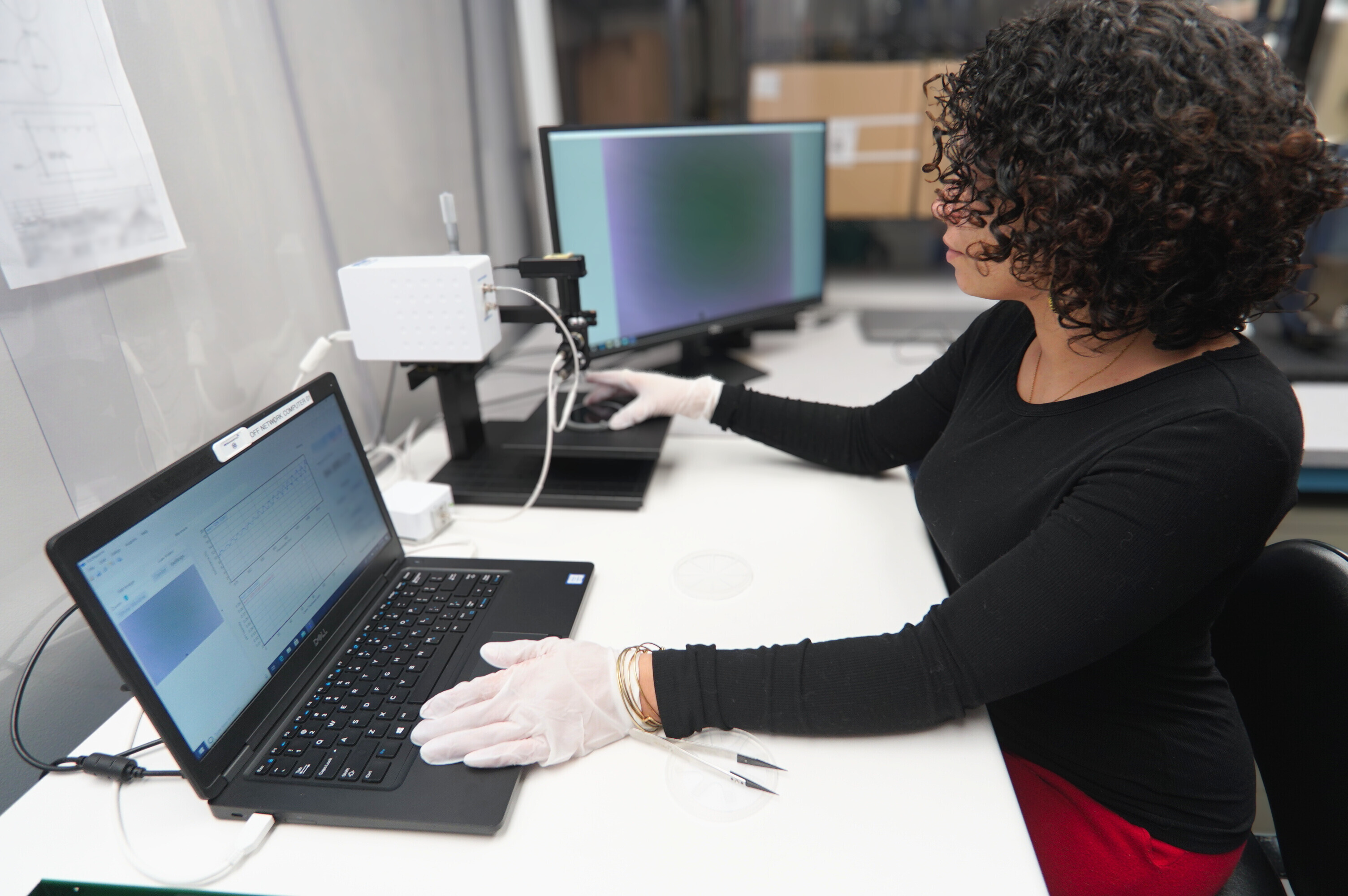 Person wearing gloves working on a laptop and microscope setup with two monitors on a white desk in a lab.