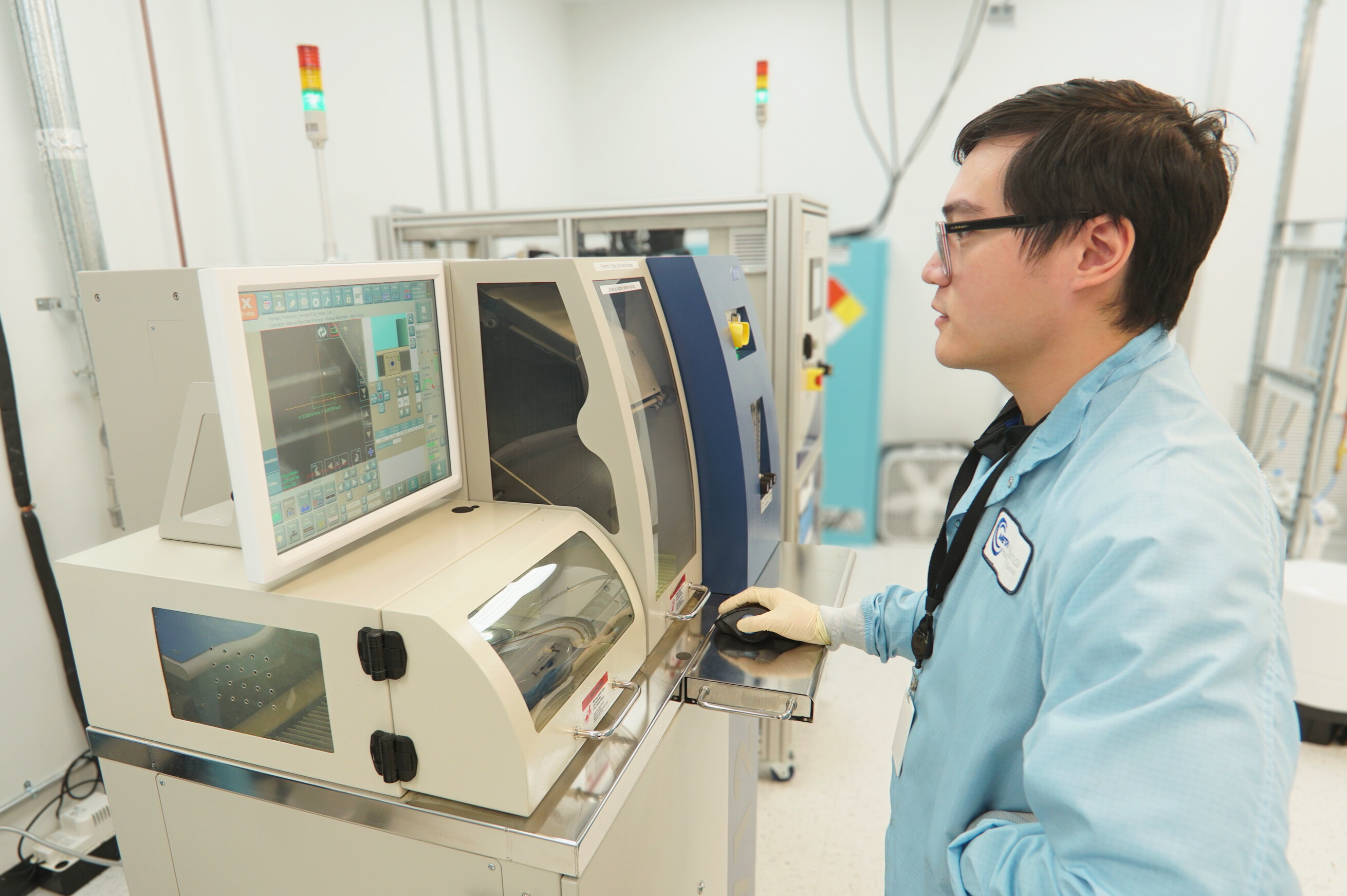 Technician in a blue lab coat operating a wafer cutting machine with a computer interface in a cleanroom.