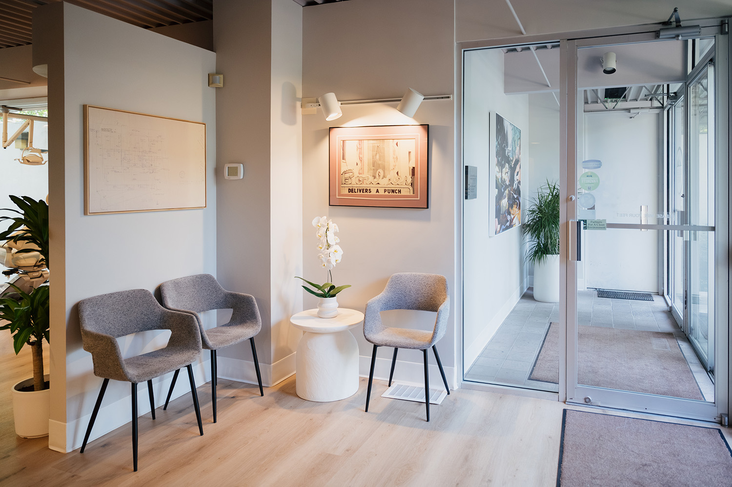 Modern waiting area with three gray chairs around a small white table with an orchid, framed artwork on the wall, and glass entrance doors.