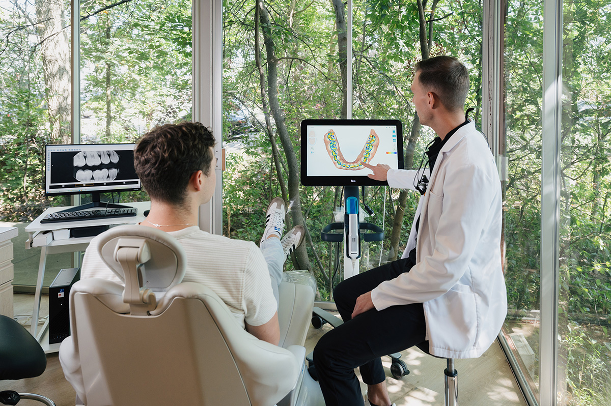 Dentist in white coat explaining dental scan on a screen to a patient sitting in a dental chair by large windows.