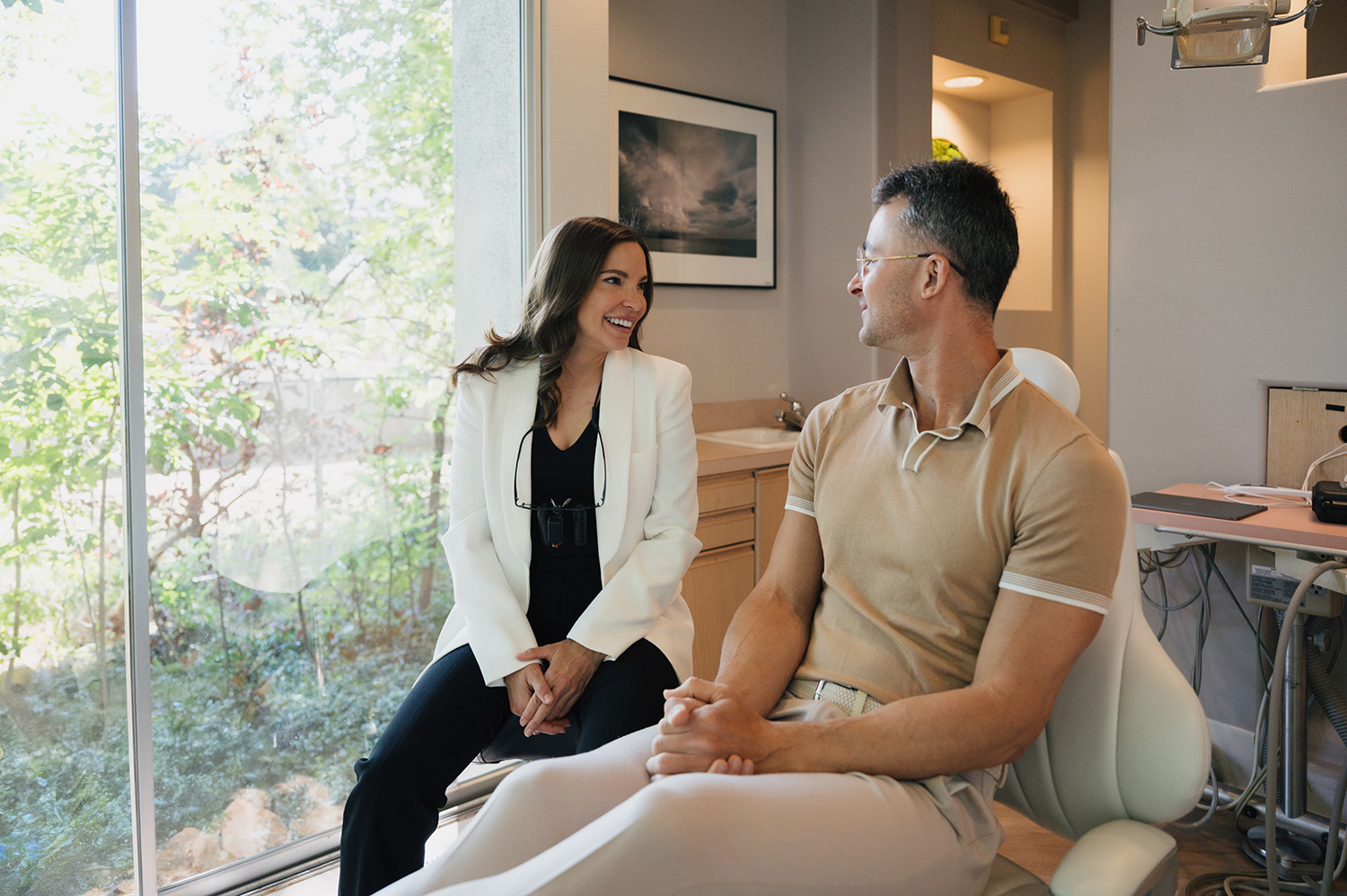 A woman in a white blazer and black outfit smiles while sitting and talking with a man in a beige polo shirt seated in a modern office with large glass windows and greenery outside.