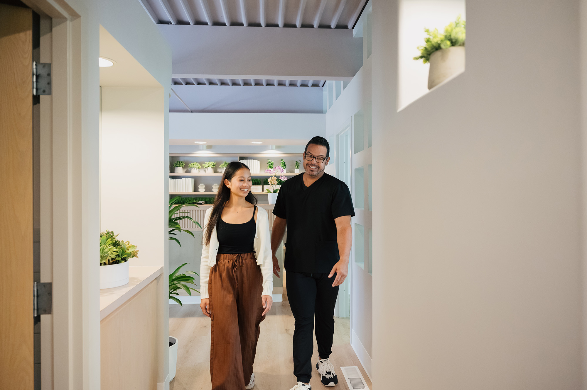 Smiling woman and man walking down a bright corridor decorated with plants.