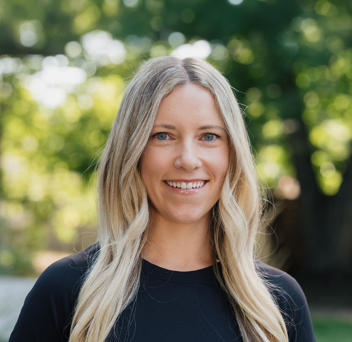 Smiling woman with long blonde hair and blue eyes wearing a black top, outdoors with blurred green trees background.